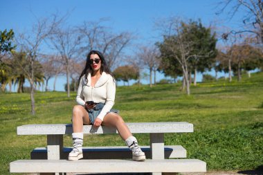 Parktaki bankta oturan genç ve güzel İspanyol esmer kadın. Kız rahat giyiniyor ve güneşli bahar gününün tadını çıkarırken cep telefonuna danışıyor.