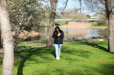 Kısa saçlı, kot pantolonlu ve siyah ceketli genç ve güzel esmer kadın soğuk bir günde yapraksız ağaçların arasında parkta yürürken kadın farklı vücut duruşları sergiliyor.
