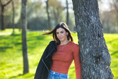 Genç ve güzel esmer kadın, kısa saçlı, kot pantolonlu, omuzlarının üzerinde turuncu gömlekli, ağaca yaslanmış, mutlu ve ormanda yürürken eğleniyor.