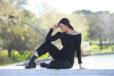 Genç ve güzel bir kadın, esmer ve siyah giyinmiş parkta bir duvarda oturuyor, arka planda sonbaharda ağaçlar ve şafakta güneş, kadın farklı vücut duruşları sergiliyor.