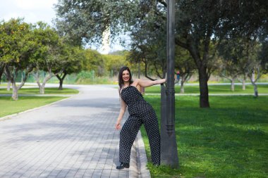 Güzel bir genç kadın, esmer ve siyah elbiseli, beyaz benekli, parkta bir sokak lambasına tutunmuş, kadın makyaj yapıyor ve kırmızı ruj sürüyor. Kadın mutlu ve gülümsüyor.