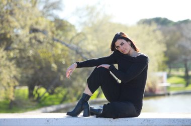 Genç ve güzel bir kadın, esmer ve siyah giyinmiş parkta bir duvarda oturuyor, arka planda sonbaharda ağaçlar ve şafakta güneş, kadın farklı vücut duruşları sergiliyor.