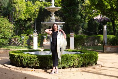 Sevilla 'dan zarif siyah bir elbise ve ipek çiçeklerle işlenmiş bej rengi ipek bir şal giymiş güzel bir İspanyol kadın parkta yürüyor.