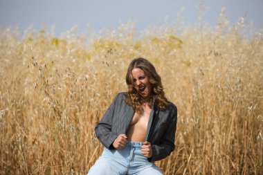 Genç, güzel sarışın kadın gri ceketli, kot pantolonlu, botlu yulaflı bir taburede oturuyor. Kadın öfke ve öfkeyle bağırıyor.