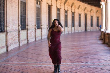 Latin kadın, genç, güzel ve şişman kıvrımlı ve uzun kıvırcık kahverengi saçlı, zarif uzun elbiseli, plaza espana 'nın kemerleri arasında yürüyor farklı pozlar ve duruşlar yapıyor.