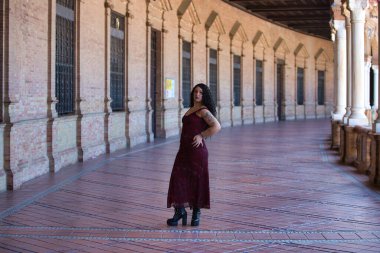 Latin kadın, genç, güzel ve şişman kıvrımlı ve uzun kıvırcık kahverengi saçlı, zarif uzun elbiseli, plaza espana 'nın kemerleri arasında yürüyor farklı pozlar ve duruşlar yapıyor.