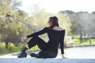 Genç ve güzel bir kadın, esmer ve siyah giyinmiş parkta bir duvarda oturuyor, arka planda sonbaharda ağaçlar ve şafakta güneş, kadın farklı vücut duruşları sergiliyor.