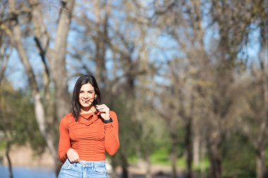 Gündelik kıyafetler ve güneş gözlüğü takmış genç ve güzel esmer kadın, sonbaharda yaprak döken ağaçlar arasında bir yol boyunca yürüyor, arka planda bir göl.