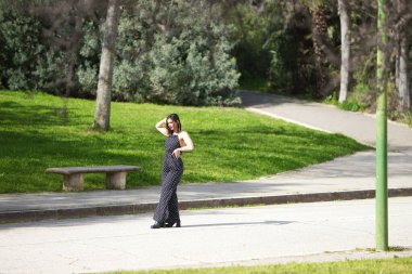 Genç ve güzel bir kadın, esmer ve siyah elbiseli, beyaz benekli, yolda fotoğraf çekimi için poz veren, makyajlı ve kırmızı rujlu bir kadın.