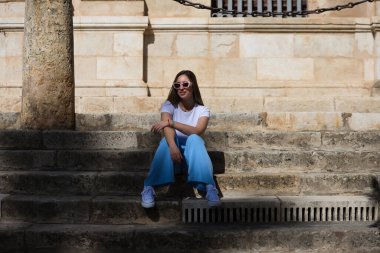 Kot pantolon, beyaz tişört ve güneş gözlüğü giymiş genç ve güzel bir kadın Sevilla 'nın merkezindeki eski bir binanın merdivenlerinde oturuyor. Kadın mutlu ve vücut duruşu yaparken eğleniyor.
