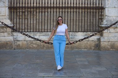 Kot pantolon, beyaz tişört ve güneş gözlüğü giymiş güzel genç bir kadın Seville 'deki gotik katedrali çevreleyen zincirlerde oturuyor. Kadın şehre yaptığı ziyaretten zevk alıyor. Turizm konsepti