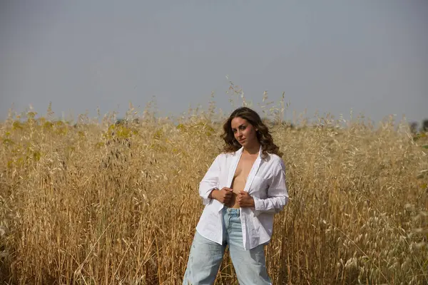 stock image pretty young blonde woman with light eyes is dressed in white shirt, jeans, boots standing in a field planted with oats. The farmer is posing for the photo, in the background the blue sky