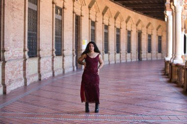 Latin kadın, genç, güzel ve şişman kıvrımlı ve uzun kıvırcık kahverengi saçlı, zarif uzun elbiseli, plaza espana 'nın kemerleri arasında yürüyor farklı pozlar ve duruşlar yapıyor.