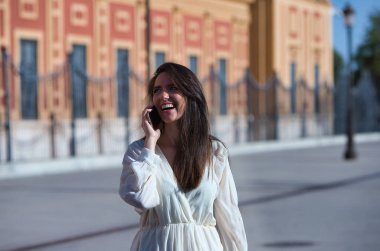 Genç ve güzel Latin kadın cep telefonuyla konuşuyor ve eğleniyor ve yüksek sesle gülümsüyor. Kadın caddede ve şehrin içinde yürüyor. Yeni teknolojiler kavramı