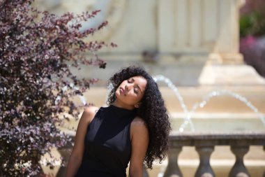 Büyük kahverengi gözleri, kıvırcık saçları ve kısa siyah elbisesi olan genç ve güzel Latin bir kadın parktaki bahçe parmaklıklarında dinleniyor. Kadın huzur içinde ve güneşlenmek için gözlerini kapatıyor.