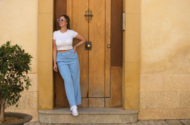 Kot pantolonlu, beyaz tişörtlü ve güneş gözlüklü genç, güzel bir kadın binanın girişine yaslanıyor. Kadın mutlu ve farklı vücut hareketleri yaparken eğleniyor.