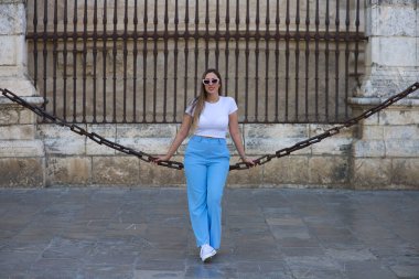 Kot pantolon, beyaz tişört ve güneş gözlüğü giymiş güzel genç bir kadın Seville 'deki gotik katedrali çevreleyen zincirlerde oturuyor. Kadın şehre yaptığı ziyaretten zevk alıyor. Turizm konsepti