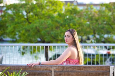 Genç, güzel, sarışın İspanyol kadın sırtı dönük bir bankta dinleniyor, kadın çiçekli ve bitki motifli bir tulum giyiyor.