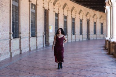 Latin kadın, genç, güzel ve şişman kıvrımlı ve uzun kıvırcık kahverengi saçlı, zarif uzun elbiseli, plaza espana 'nın kemerleri arasında yürüyor farklı pozlar ve duruşlar yapıyor.