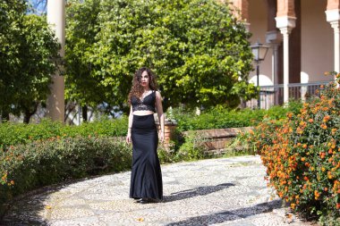 Beautiful woman with long curly hair, walks in a park in Seville, Spain. Dressed in long black skirt and black top. The woman is a flamenco dancer and takes advantage of her break to visit the city clipart