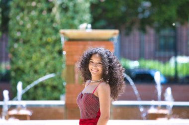 Büyük gözlü, esmer, kıvırcık saçlı, kırmızı elbiseli, parktaki ağaçların arasında yürüyen ve farklı yüz ifadeleri takınan güzel Latin kadın portresi. Moda konsepti