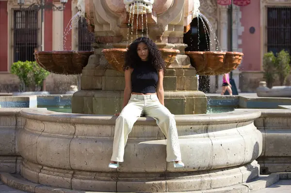 stock image beautiful young latin woman with black and dark eyes and long curly hair rests sitting on a pink and white marble fountain in front of the cathedral of Seville, andalusia, Spain