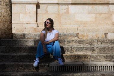 Kot pantolon, beyaz tişört ve güneş gözlüğü giymiş genç ve güzel bir kadın Sevilla 'nın merkezindeki eski bir binanın merdivenlerinde oturuyor. Kadın mutlu ve vücut duruşu yaparken eğleniyor.