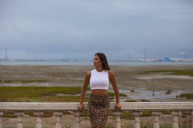 Latin kadın, genç ve güzel beyaz gömlek ve kaplan desenli etek giymiş taş korkuluklara yaslanmış. Arka planda tuz madeni ve ufukta Cadiz şehri var.