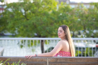 Genç, güzel, sarışın İspanyol kadın sırtı dönük bir bankta dinleniyor, kadın çiçekli ve bitki motifli bir tulum giyiyor.