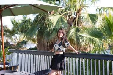 Tatilde olan genç ve güzel Latin kadın demir korkuluklara, arka plandaki ağaçlara ve Seville 'deki nehrin diğer kıyısına yaslanıyor. Kadın farklı ifadeler kullanıyor.