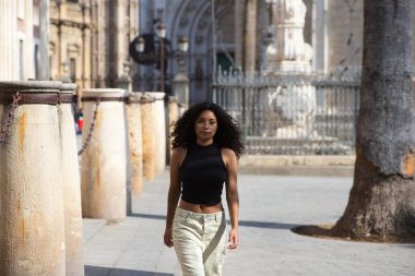 Koyu renk gözlü, uzun kıvırcık saçlı güzel bir Latin kadın Sevilla 'nın merkezinde yürüyor. Arka planda katolik tapınma katedralini ve Sevilla' nın gotik tarzını görebilirsiniz.