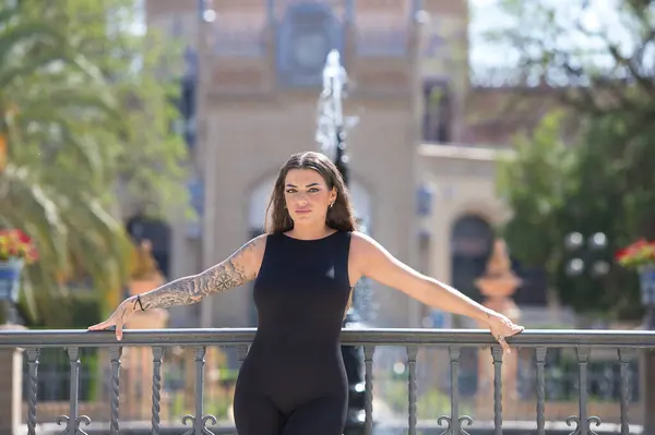 Siyah likra tulum giymiş genç ve güzel esmer kadın işlenmiş demir parmaklıklara yaslanıyor. Arka planda, Endülüs, Sevilla Sanat ve Gümrük Müzesi var.