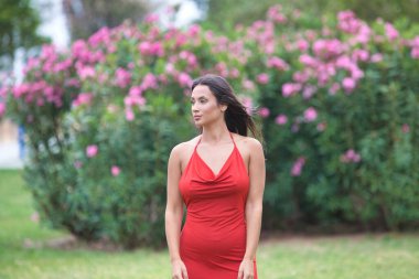 Uzun saçlı, yeşil gözlü, çok zarif kırmızı elbiseli genç ve güzel İspanyol kadın parkta çimlerin üzerinde manken gibi yürüyor ve farklı vücut duruşları sergiliyor.