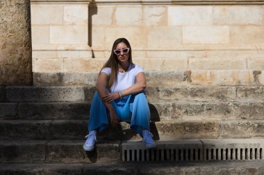 Kot pantolon, beyaz tişört ve güneş gözlüğü giymiş genç ve güzel bir kadın Sevilla 'nın merkezindeki eski bir binanın merdivenlerinde oturuyor. Kadın mutlu ve vücut duruşu yaparken eğleniyor.