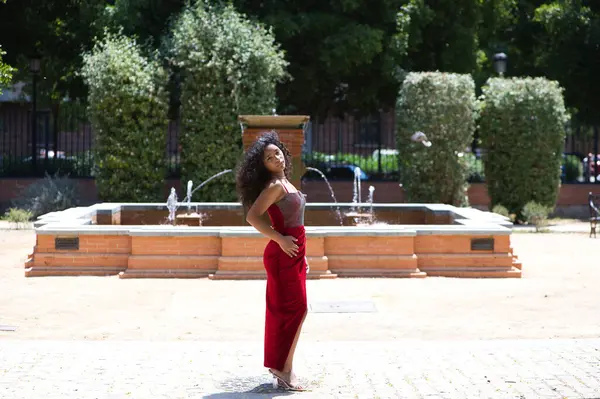 Büyük gözlü, kıvırcık saçlı, kırmızı elbiseli güzel Latin bir kadın fotoğraf çekimi için parkta poz veriyor. Arka planda bir çeşme var. Moda ve güzellik konsepti