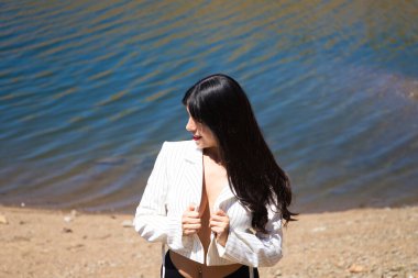 Kırmızı dudaklı, beyaz gömlekli, siyah şortlu, gömleğini elleriyle tutan güzel, esmer bir kadının portresi. Arka planda kristal berrak mavi suyu olan bir göl.