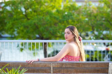 Genç, güzel, sarışın İspanyol kadın sırtı dönük bir bankta dinleniyor, kadın çiçekli ve bitki motifli bir tulum giyiyor.