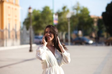 Genç ve güzel Latin kadın cep telefonuyla konuşuyor ve eğleniyor ve yüksek sesle gülümsüyor. Kadın caddede ve şehrin içinde yürüyor. Yeni teknolojiler kavramı