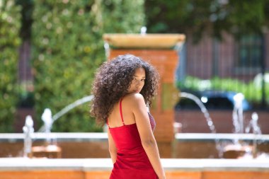 Büyük gözlü, esmer, kıvırcık saçlı, kırmızı elbiseli, parktaki ağaçların arasında yürüyen ve farklı yüz ifadeleri takınan güzel Latin kadın portresi. Moda konsepti