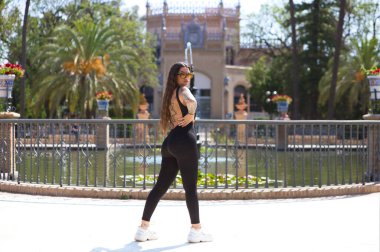 Siyah likra tulumlu genç esmer kadın Seville 'de bir parkta yapay göle poz veriyor. Arka planda Arap tarzı sanat ve gelenek müzesi var.
