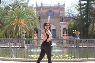Siyah likra tulumlu genç esmer kadın Seville 'de bir parkta yapay göle poz veriyor. Arka planda Arap tarzı sanat ve gelenek müzesi var.