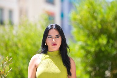 Genç İspanyol kadın portresi, yeşil elbiseli güzel esmer. Arka planda şehir parkındaki ağaçlar ve bitki örtüsü var. Kadın ciddi.