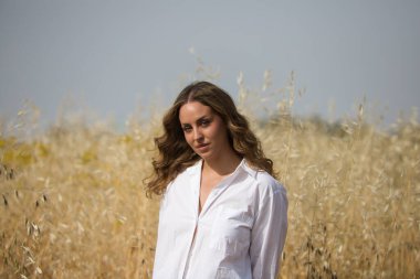 Açık renk gözlü güzel sarışın kadın beyaz gömlek, kot pantolon, botlar giymiş yulaflı bir tarlada dikiliyor. Çiftçi fotoğraf için poz veriyor, arka planda mavi gökyüzü