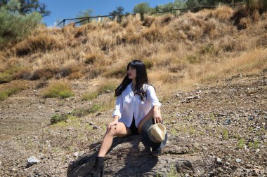 Genç, esmer, genç ve güzel Latin kadın beyaz gömlek ve siyah şort giymiş ve saman şapkalı tarlada oturuyor. Kadın farklı vücut ve yüz duruşları yapıyor.