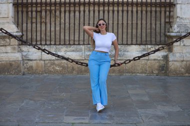 Kot pantolon, beyaz tişört ve güneş gözlüğü giymiş güzel genç bir kadın Seville 'deki gotik katedrali çevreleyen zincirlerde oturuyor. Kadın şehre yaptığı ziyaretten zevk alıyor. Turizm konsepti