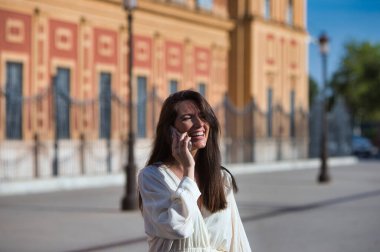 Genç ve güzel Latin kadın cep telefonuyla konuşuyor ve eğleniyor ve yüksek sesle gülümsüyor. Kadın caddede ve şehrin içinde yürüyor. Yeni teknolojiler kavramı
