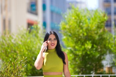 Beautiful young Spanish brunette woman in a green dress talking on the phone standing in a city park. The woman follows the conversation attentively. Concept of mobile telephony and technology clipart