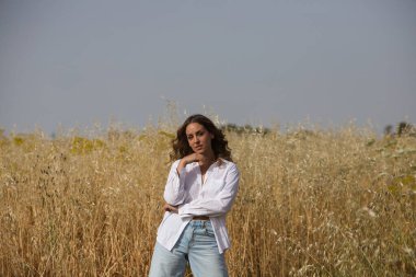 Açık renk gözlü güzel sarışın kadın beyaz gömlek, kot pantolon, botlar giymiş yulaflı bir tarlada dikiliyor. Çiftçi fotoğraf için poz veriyor, arka planda mavi gökyüzü