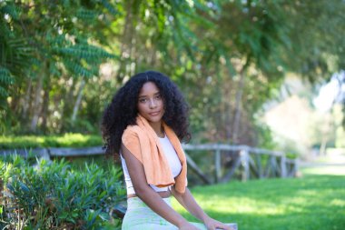 Genç, güzel, Latin, kıvırcık saçlı, spor kıyafetleri giyen ve terini kurutmak için boynuna havlu takan esmer kadın ahşap bir çitte oturuyor. Spor ve sağlık kavramı