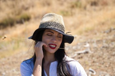 Beyaz gömlek ve hasır şapka giymiş, güzel, genç, esmer bir kadının portresi. Arka planda ağaçlar ve bitki örtüsü var.